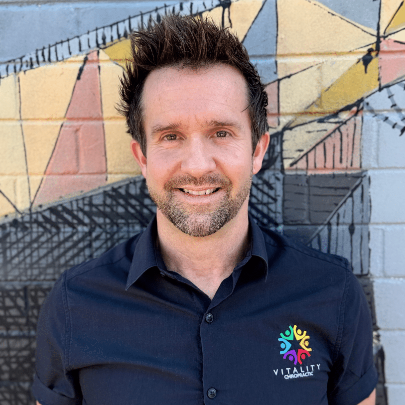 A smiling man with short, spiked brown hair stands in front of a colorful geometric mural, wearing a navy shirt with a multicolored logo and the text "Vitality Chiropractic" on it.