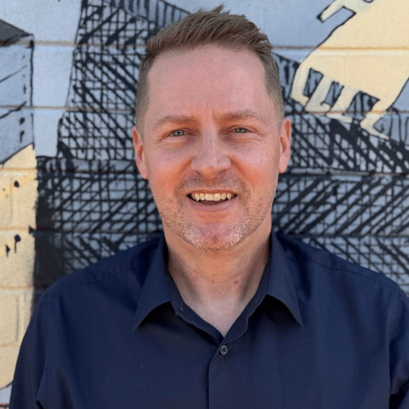 A smiling man with short light hair wearing a dark shirt stands in front of a mural painted on a brick wall. The mural features bold black lines and shapes on a light background.